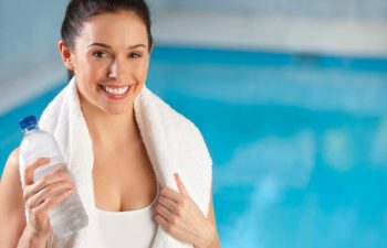 A fit woman with a bottle of water and a towel by a swimming pool.