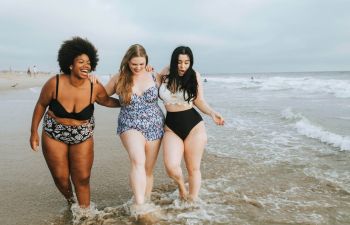 Three overweight young woman in swimming suits walking along the beach.