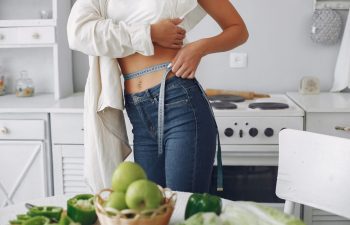 Slim sporty woman measuring her waist standing at a kitchen table full of fruit and vegetables.