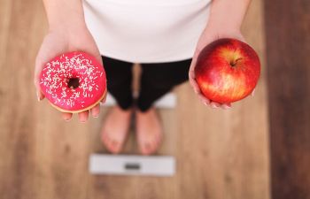 A woman standing on weighing scales with a donut and an apple in her hands.