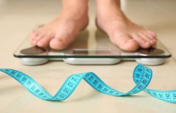 A blue measuring tape next to a person after weight loss surgery standing on weighing scales.