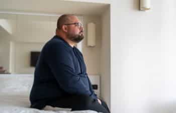 plus sized man sitting on bed in hotel and looking out window