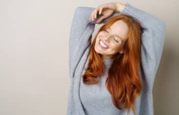 Woman with long red hair in a gray sweater smiling with her eyes closed and arms raised over her head against a plain background.